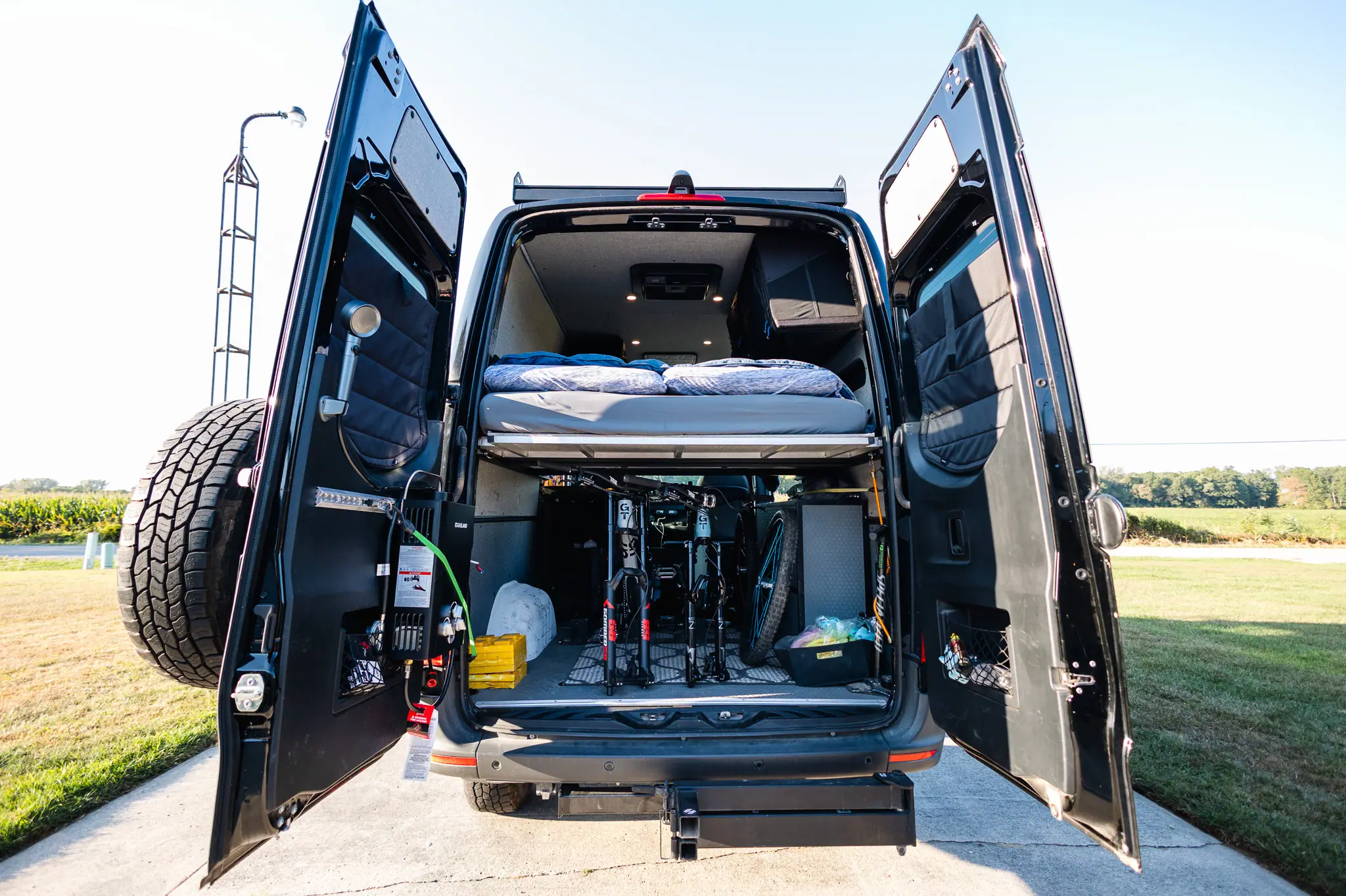 Rear doors open on a black Sprinter camper van, showing the bike garage under the bed and an outdoor shower