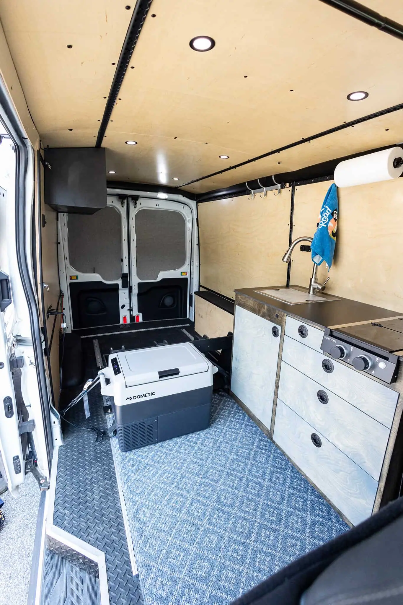 Ford Transit mid-roof van interior with plywood walls, L-track rails, a galley with sink and stove, and a fridge